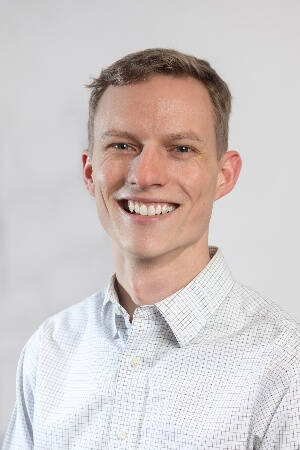 A man in an official shirt smiles at a camera in front of a gray background.